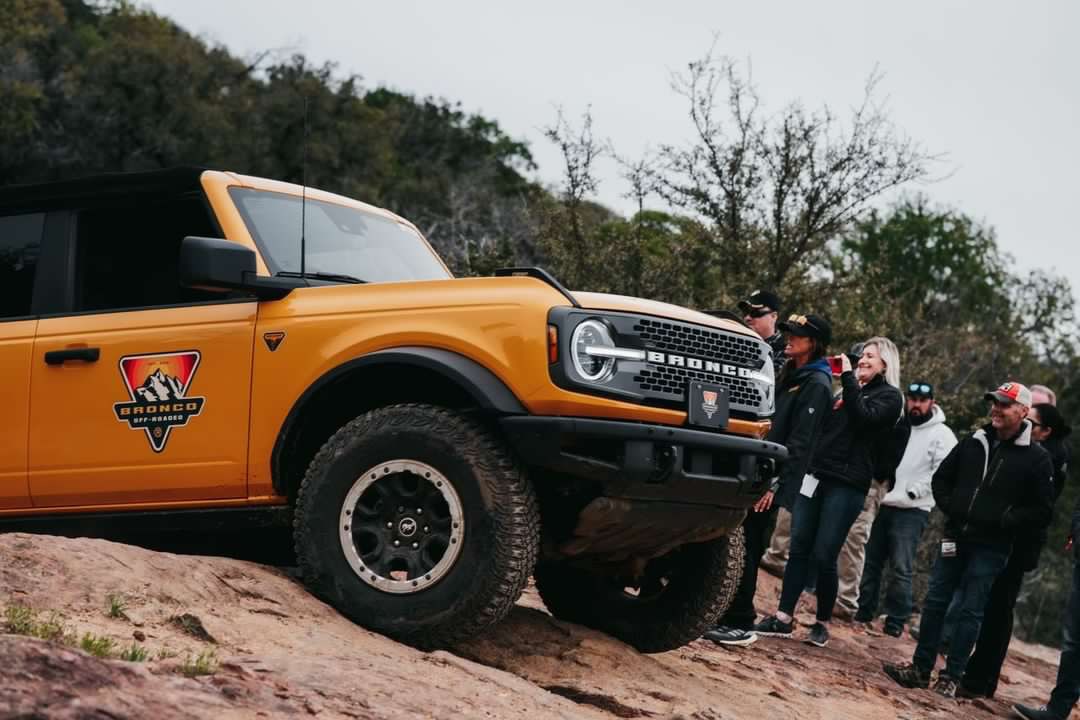 A small crowd looks at an orange-yellow Bronco parked on rocky terrain with a forested background. The Bronco has a "Bronco Off-Roadeo" decal on the side.